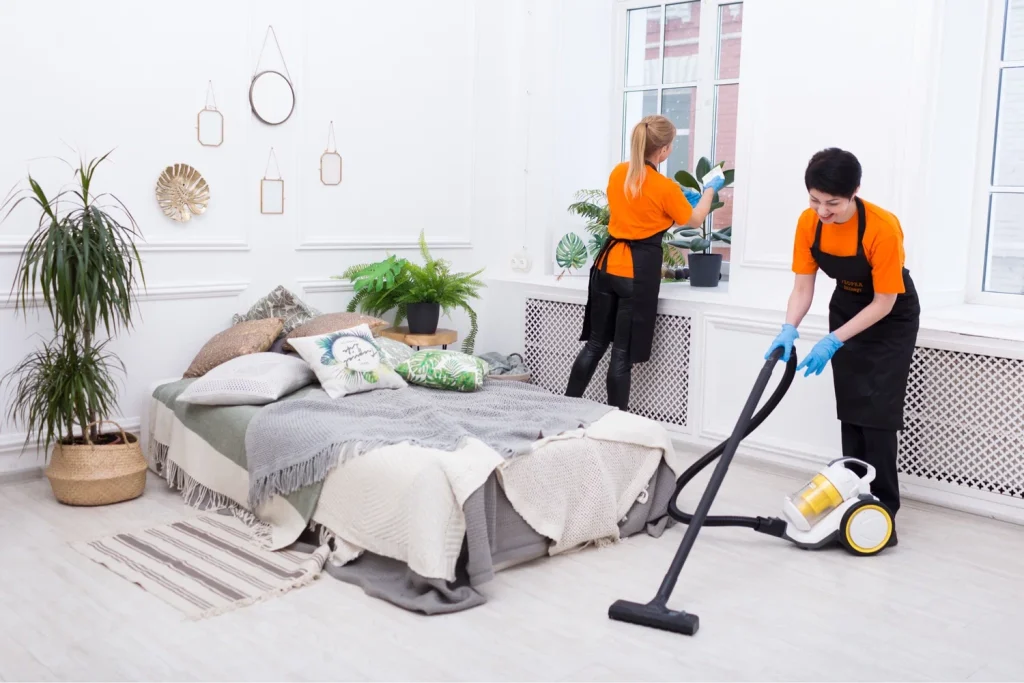 Two professional cleaners tidying a bright bedroom—one vacuuming the floor, the other cleaning the window area.