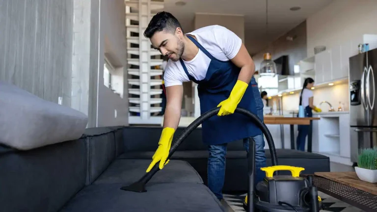 Professional cleaner vacuuming gray sofa with yellow gloves in modern home while team cleans kitchen background